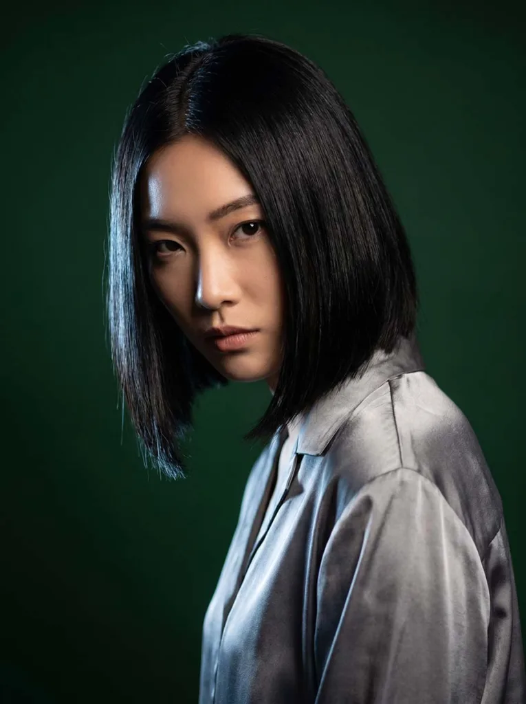 asymmetrical glass bob on East Asian woman, deep black hair, silver-grey silk blouse, forest green background, Dutch angle