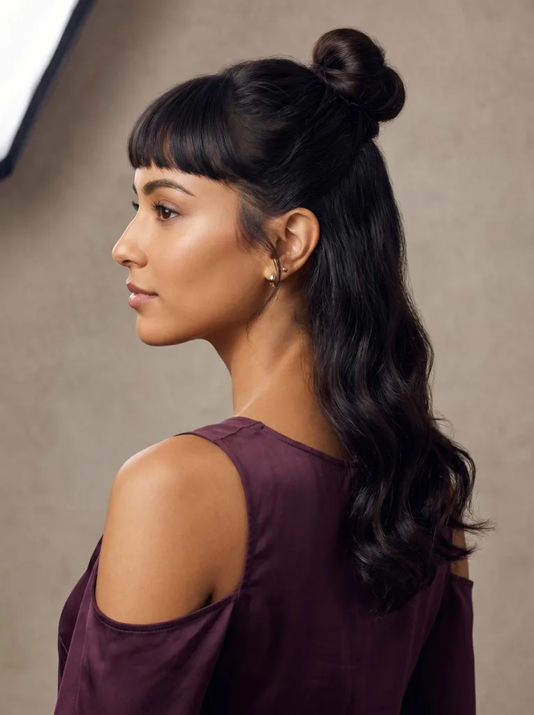 Gorgeous model with baby bangs and a chic high half-up bun, olive skin, deep plum top, elevated rear three-quarter editorial portrait