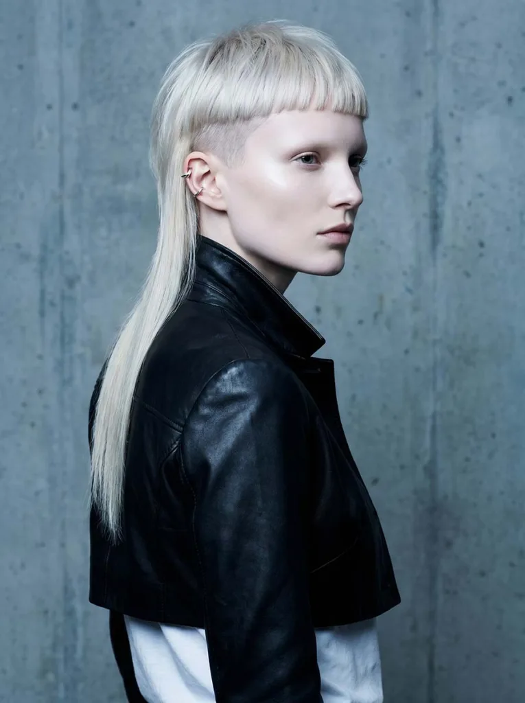 Edgy beautiful model with a platinum baby bang mullet, cool pale skin, black leather jacket, rear three-quarter urban editorial portrait