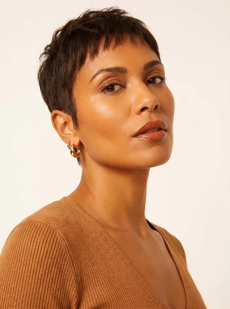 Stunning model with a chic baby bang pixie cut, warm amber glowing skin, overhead editorial portrait on warm white background