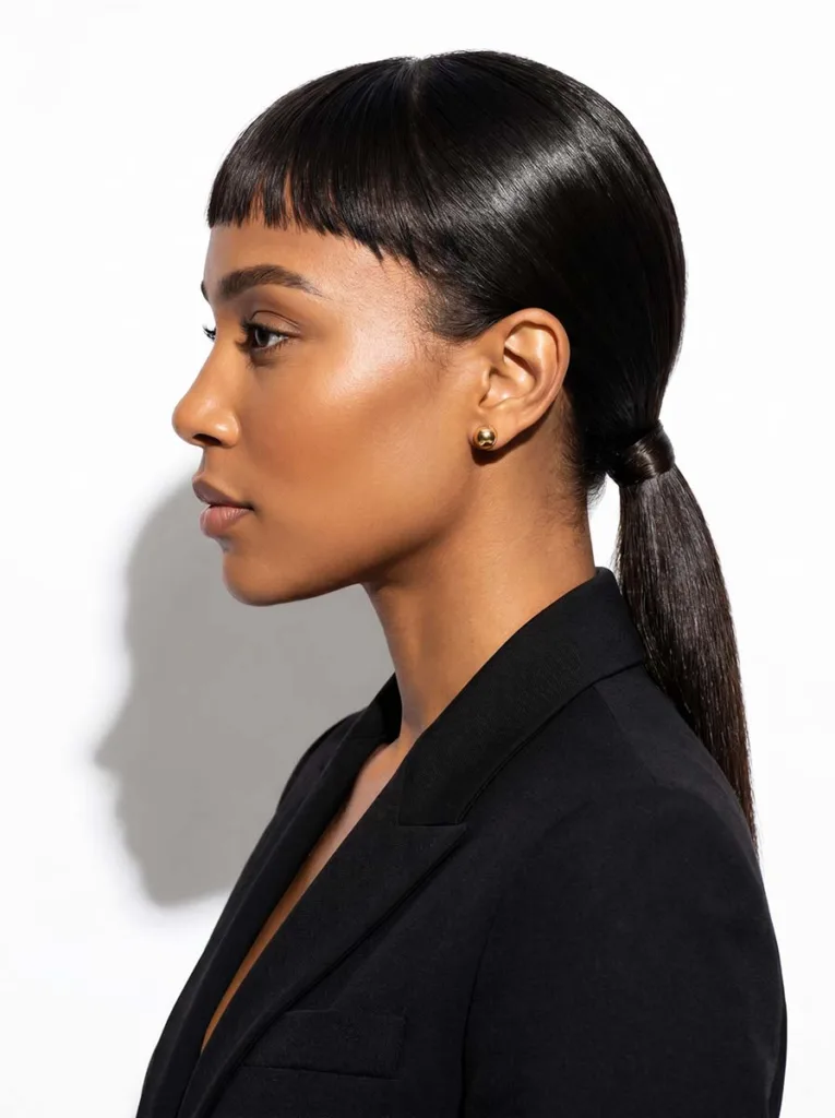 Stunning model with baby bangs and a flawless sleek low ponytail, radiant deep brown skin, black blazer, sharp side profile on white seamless