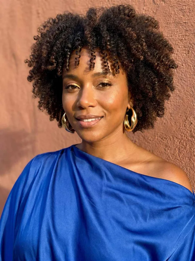 Stunning model with sculptural coily baby bangs and full natural afro, radiant deep brown skin, cobalt blue off-shoulder top, low-angle editorial portrait