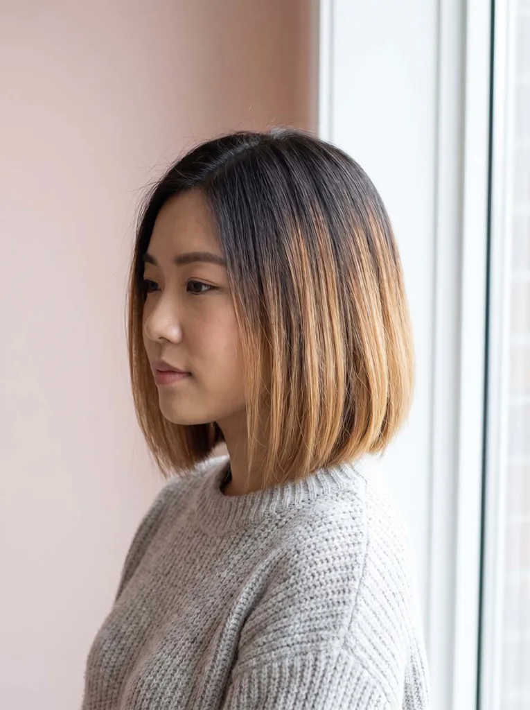 balayage glass bob on East Asian woman, dark roots to caramel ends, grey knit sweater, blush pink background