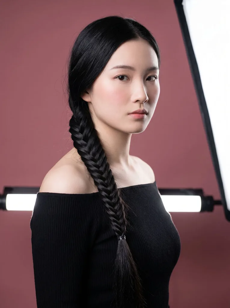 Young woman with a classic full french fishtail braid in a black off-shoulder top against a dusty rose background