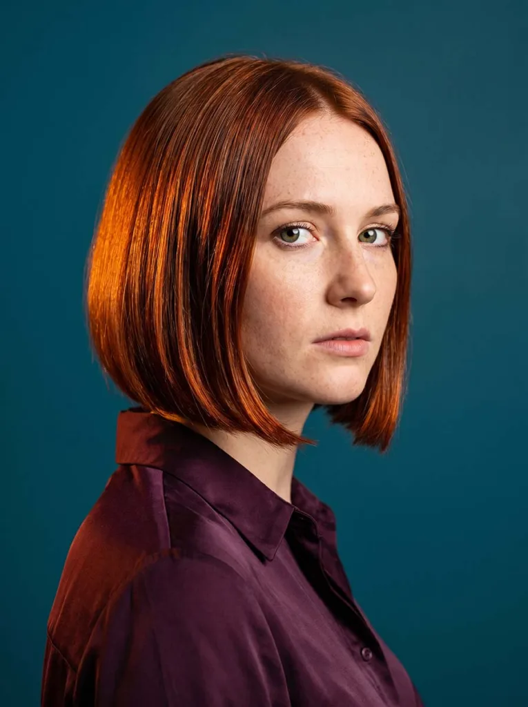 copper auburn glass bob on white woman with green eyes, deep plum blouse, teal background, warm amber side lighting