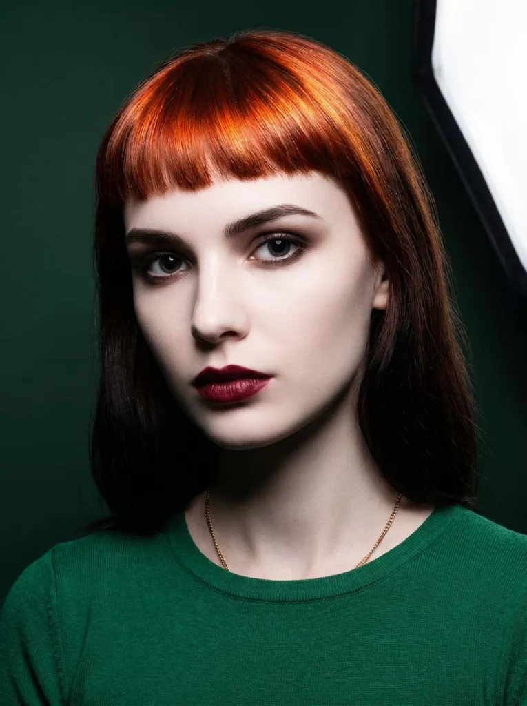 Striking beautiful model with glowing copper-red baby bangs, flawless porcelain skin, emerald top, dramatic close portrait on forest green