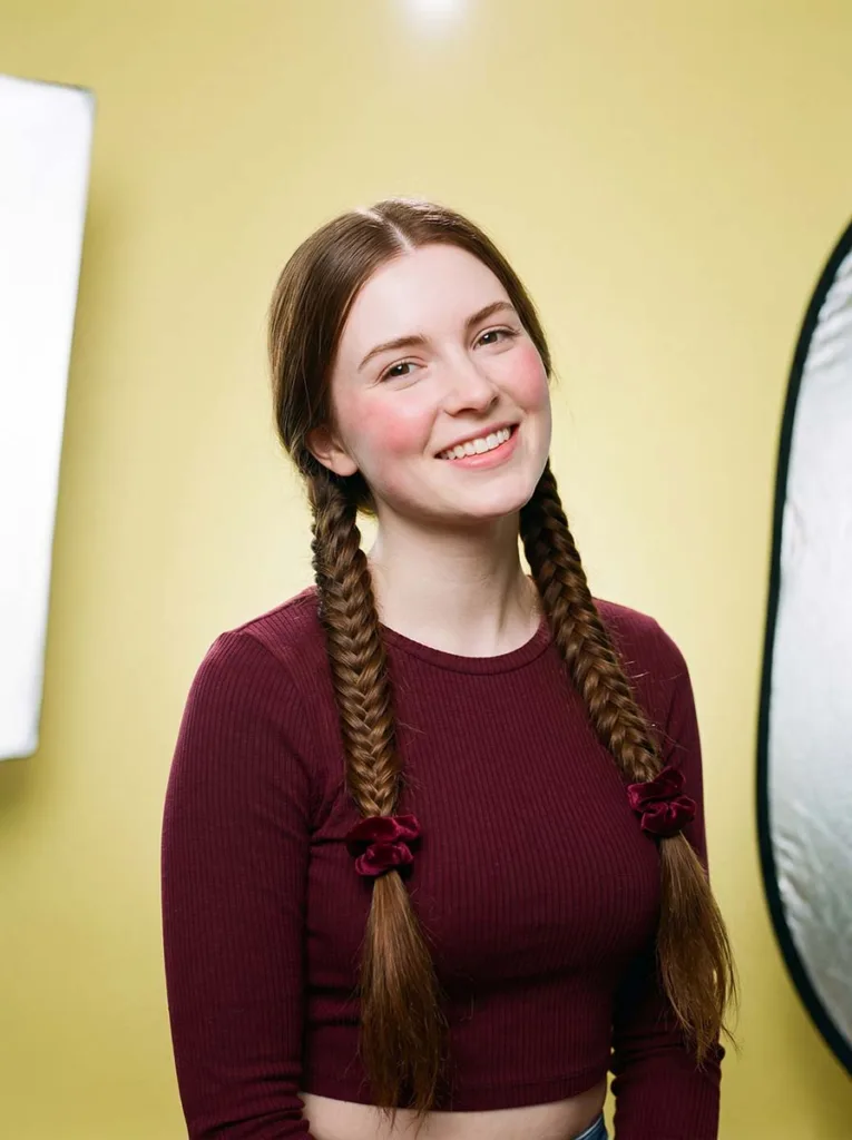 Smiling 22-year-old with double french fishtail braids and burgundy scrunchies against a pale yellow background