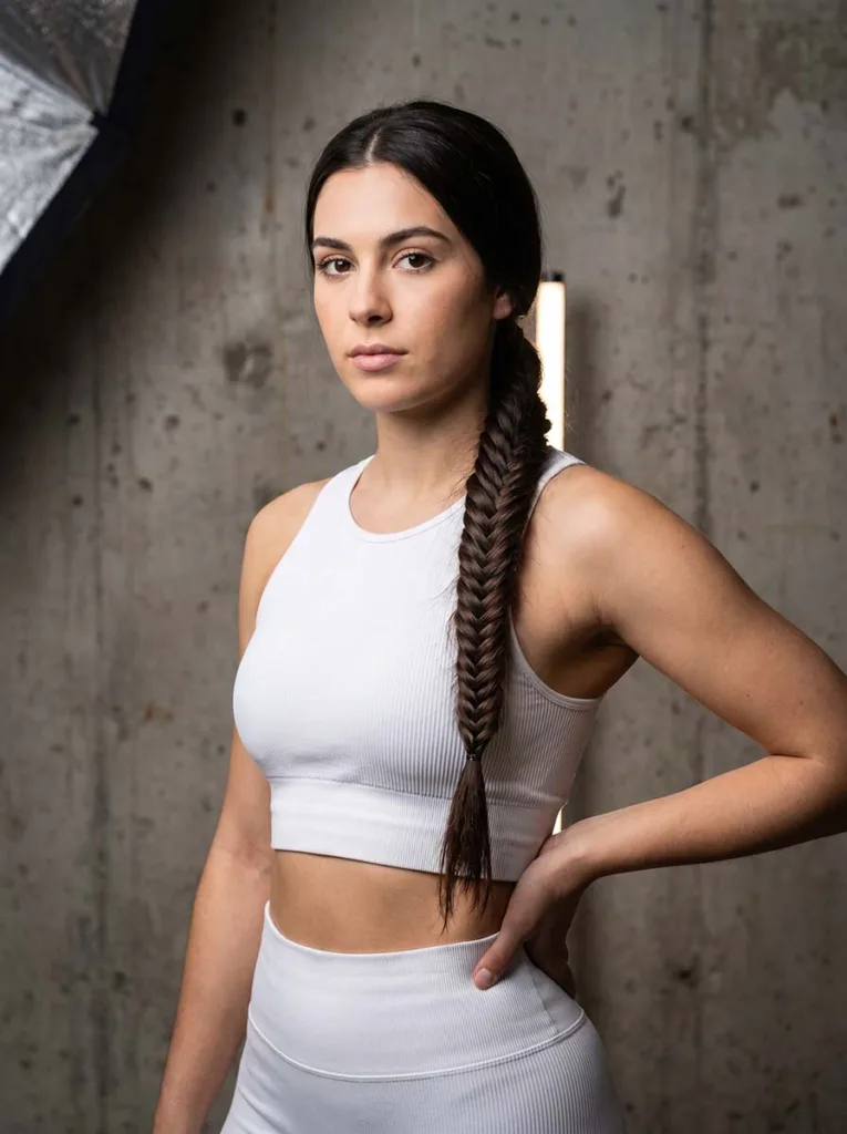 Young woman with a fishtail braid ponytail wearing a white athletic set against a concrete grey background
