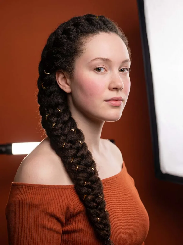 3/4 front view of a 22-year-old with a beaded french fishtail braid draped over her shoulder, gold rings glinting against natural 4C hair