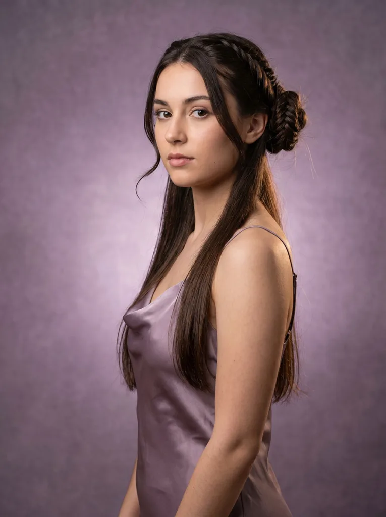 22-year-old with a french fishtail half-up bun and loose dark hair below, photographed at a 3/4 front-left angle against a dusty lavender background