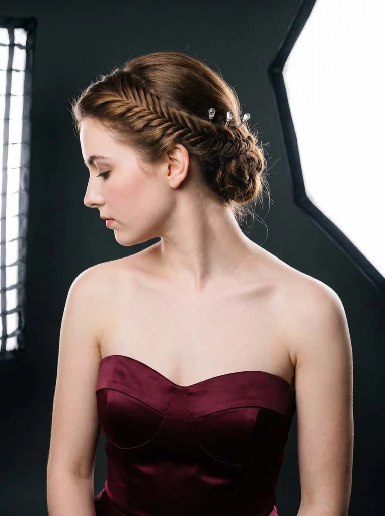 3/4 front-side view of a young woman wearing a french fishtail updo pinned at the back of the head, in a strapless burgundy gown