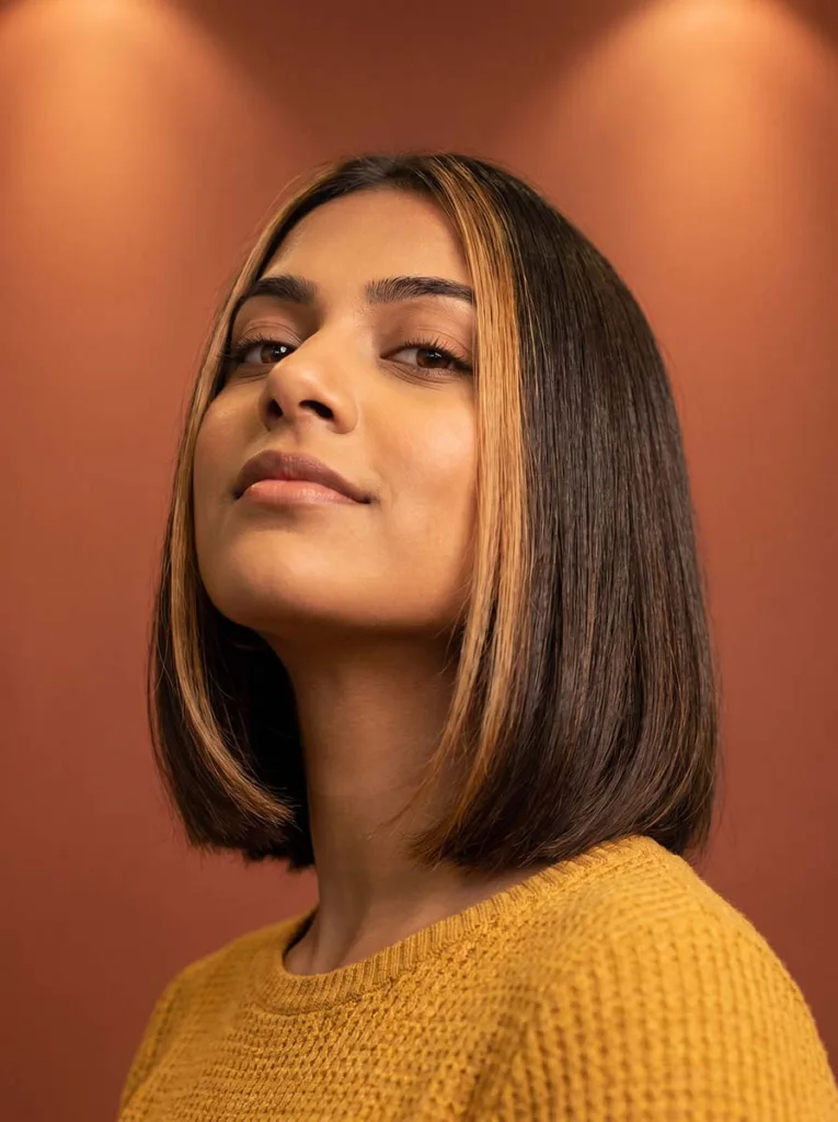 glass bob with money pieces on South Asian woman, dark base with lighter face-framing sections, mustard top, rust background