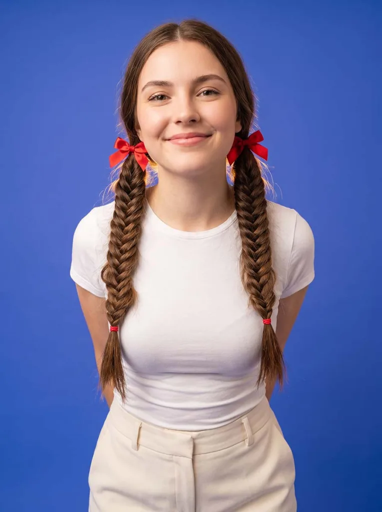 22-year-old woman with high french fishtail pigtails and red bow ties, smiling, photographed at a low angle against a bright cobalt blue background