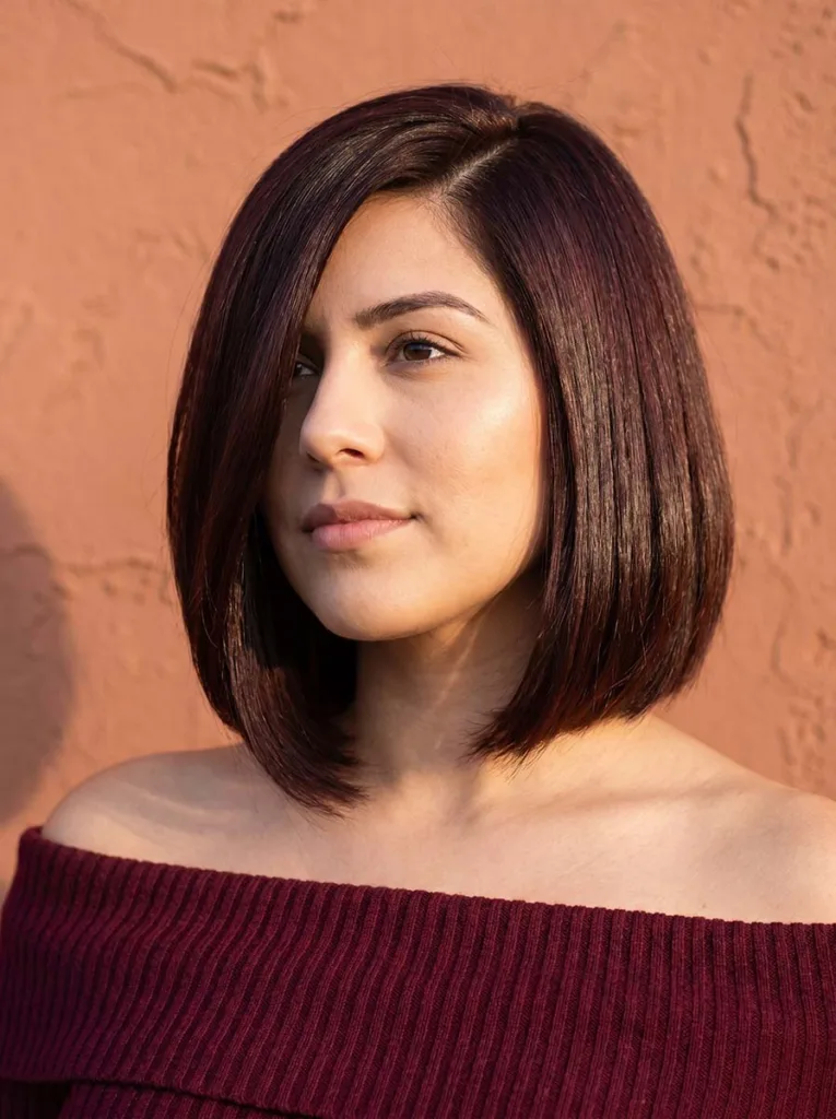 jaw-length glass bob with deep side part on Latina woman, dark mahogany hair, burgundy top, terracotta background