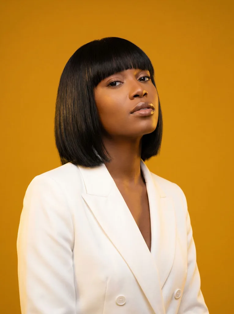 retro blunt fringe glass bob on Black woman, deep black hair, white structured blazer, mustard yellow background