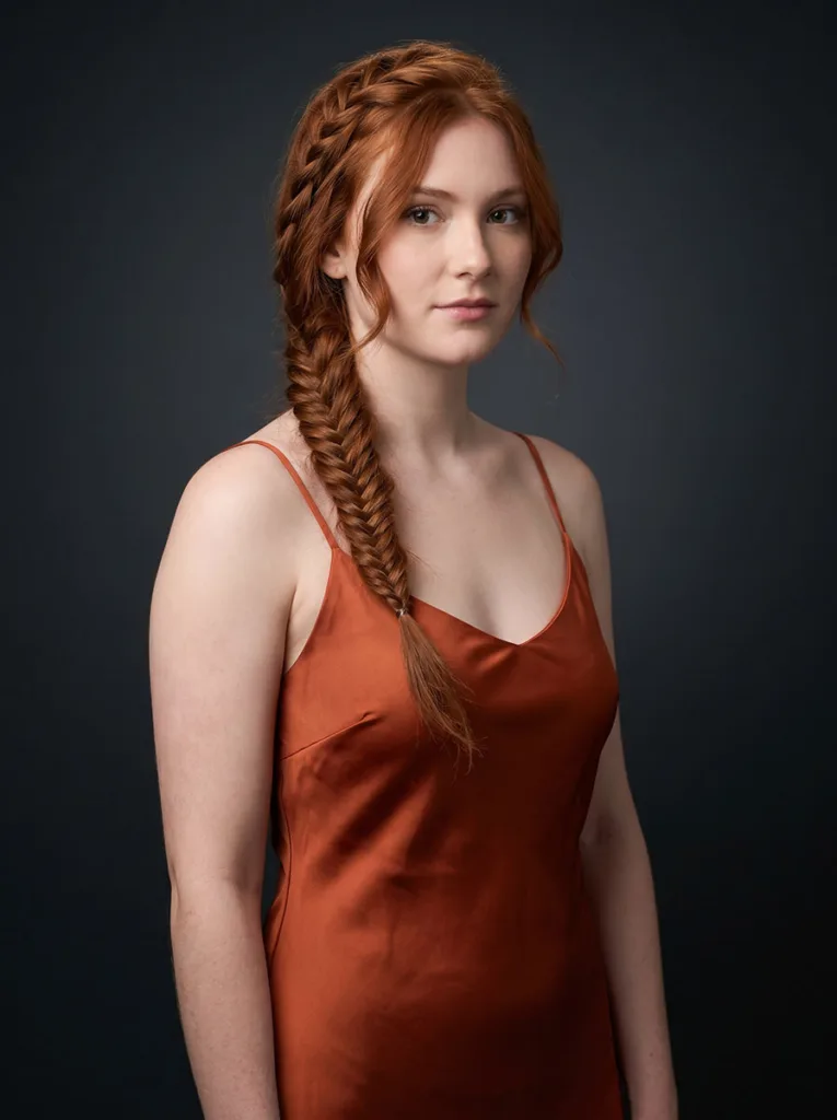 Young woman with a side french fishtail braid falling over left shoulder, wearing a rust-orange dress against a charcoal grey background