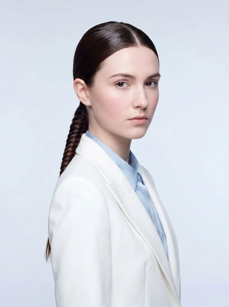 22-year-old woman with a sleek low french fishtail braid wearing a white structured blazer against a pale blue-grey background