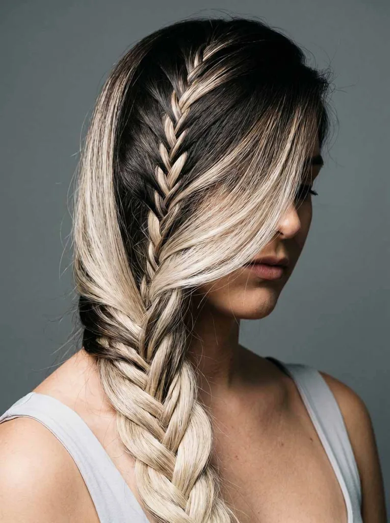 Close-up 3/4 front view of a young woman with a two-tone balayage french fishtail braid draped over her shoulder, showing the dark-to-blonde herringbone contrast
