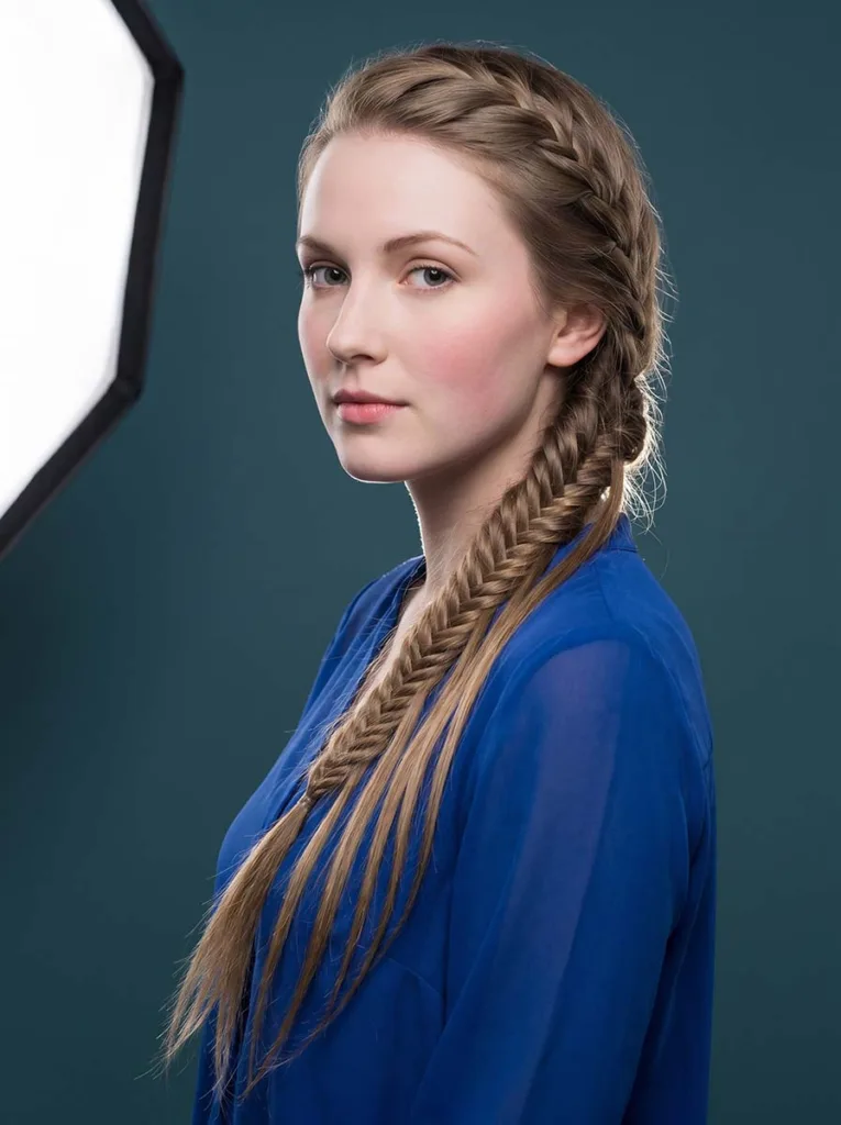 3/4 front view of a 22-year-old with a waterfall french fishtail braid, cascading strands falling forward over her shoulder against a deep teal background