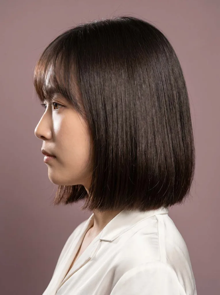 wispy fringe glass bob profile view on East Asian woman, fine dark hair, pearl-white blouse, dusty mauve background