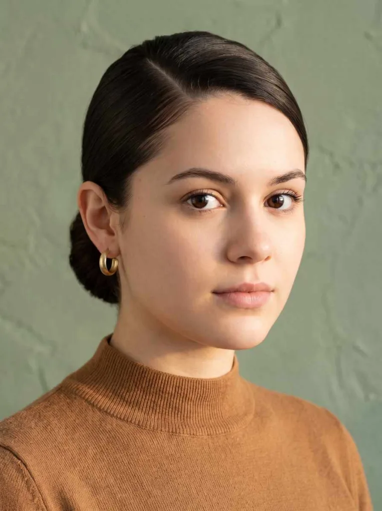 Pretty 21-year-old woman with fair skin and dark brown hair in a deep side-part sleek low bun, front-facing portrait, sage green background