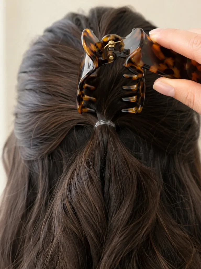 Close-up macro rear view of the back of a woman's head with dark wavy hair showing a thin clear elastic securing the half up section just before a large tortoiseshell claw clip is placed over it