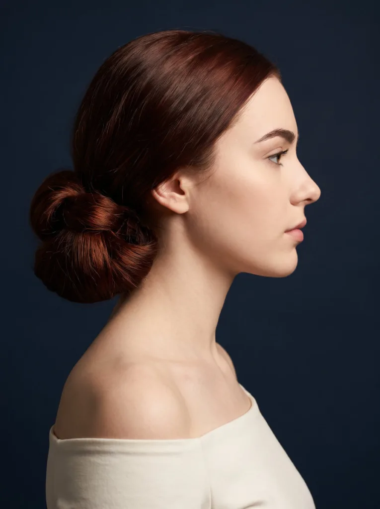 Attractive 23-year-old woman with fair skin and dark auburn hair in a knotted low bun, right side profile portrait, ivory top, navy background