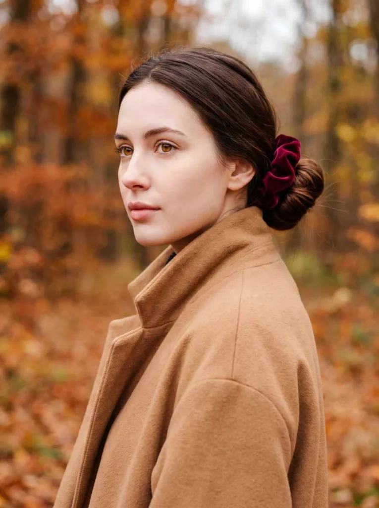 Pretty 21-year-old woman with fair porcelain skin and deep chestnut hair in a velvet scrunchie low bun, 3/4 front portrait, camel coat, autumn forest background
