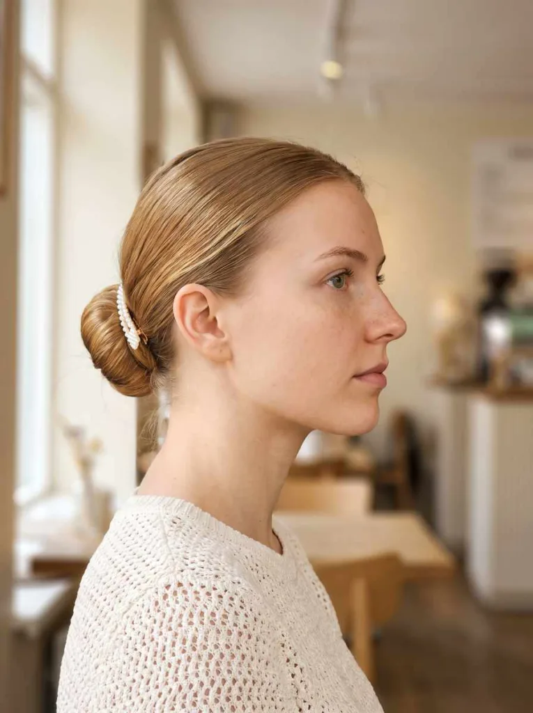 Pretty 21-year-old woman with fair Nordic skin and honey blonde hair in a sleek low bun with pearl clip, side profile portrait, café background