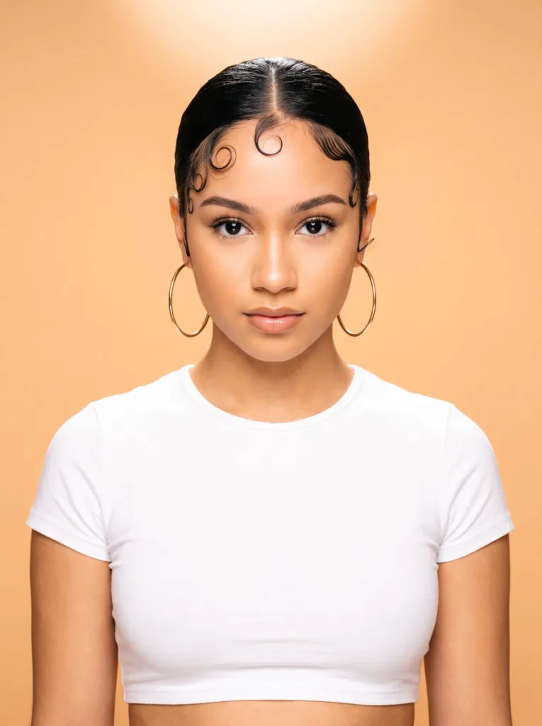 Pretty 21-year-old woman with fair warm skin and black hair with styled baby hair swirls, front-facing portrait, peach studio background