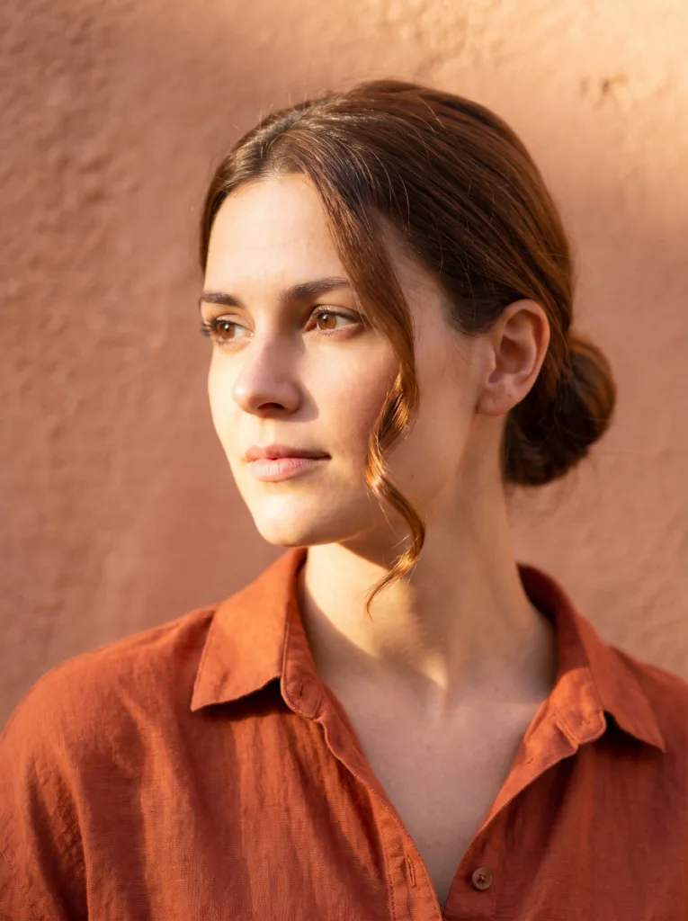 Beautiful 24-year-old woman with fair golden skin and chestnut hair in a sleek low bun with curled face-framing tendrils, 3/4 front portrait, terracotta wall background