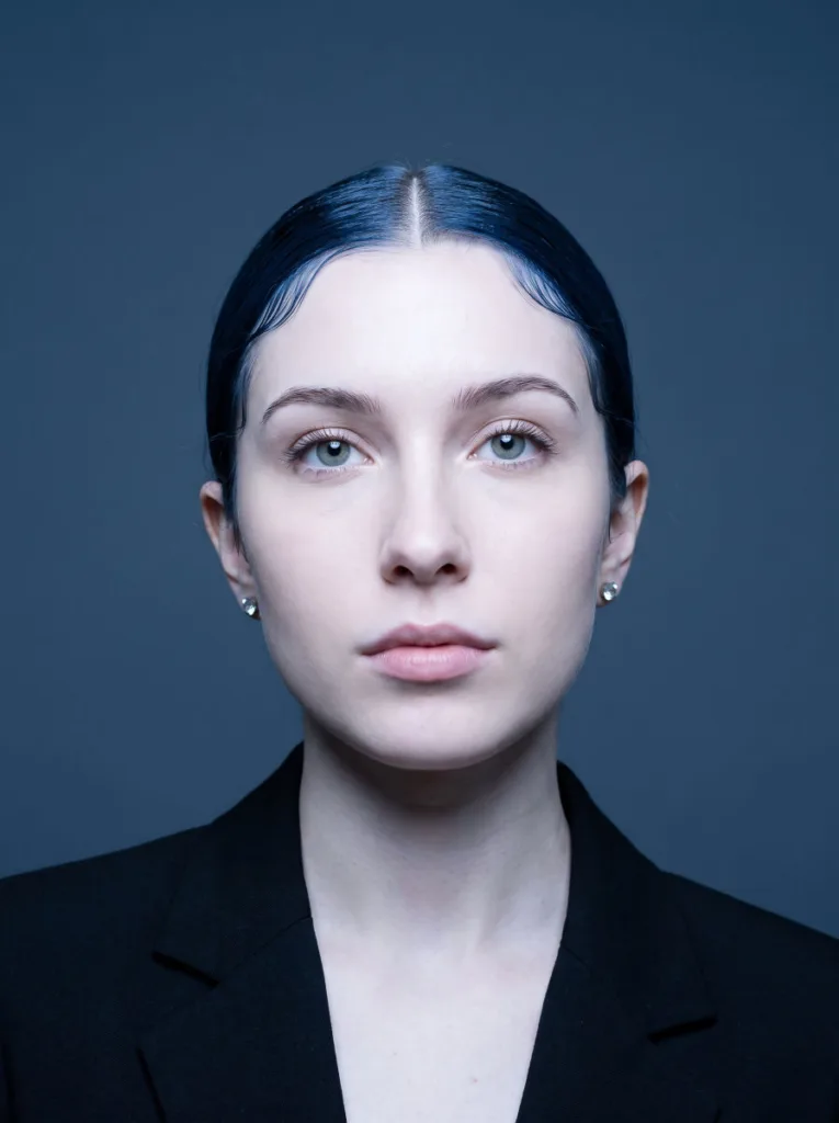 Stunning 22-year-old woman with fair ivory skin and blue-black hair in a sleek low bun with flat laid edges, front-facing editorial portrait, slate blue background