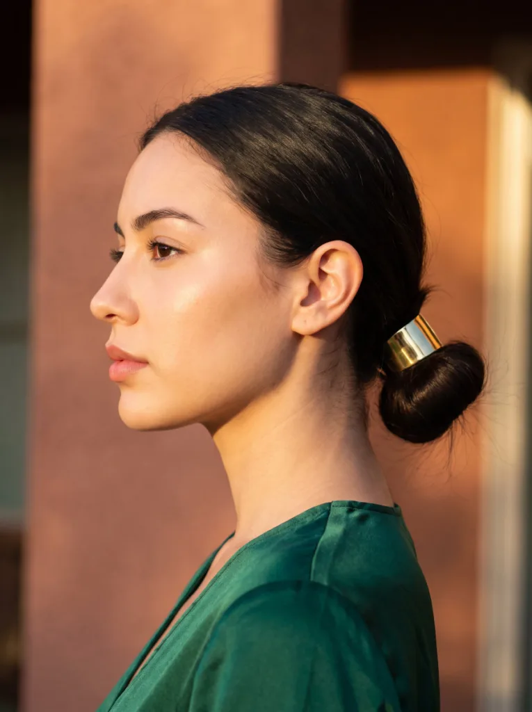 Attractive 24-year-old woman with fair golden skin and dark brown hair in a sleek low bun with gold hair cuff, left side profile portrait, emerald green top, terracotta wall background