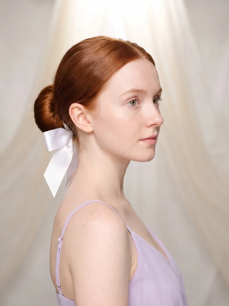 Pretty 21-year-old woman with fair freckled skin and auburn hair in a sleek low bun with white satin ribbon bow, 3/4 side portrait, lavender dress, cream background