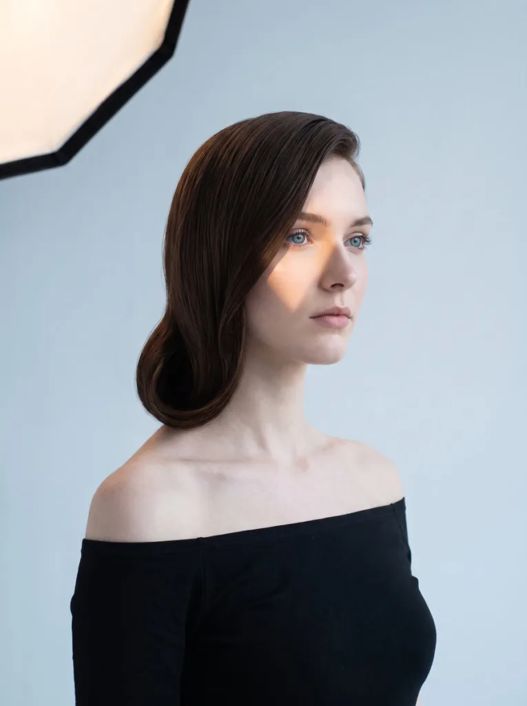 Beautiful 24-year-old woman with porcelain skin and dark chocolate hair in a sleek side swoop low bun, 3/4 front portrait, pale blue-grey background