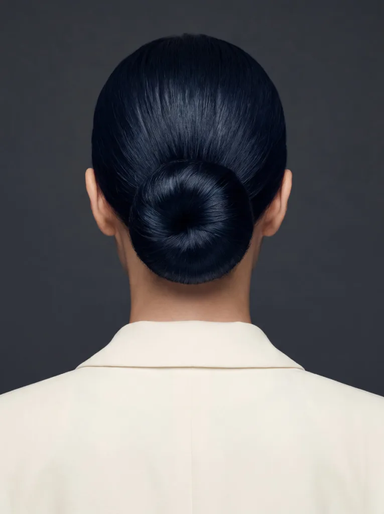 Elegant 22-year-old woman with fair porcelain skin and blue-black hair in an ultra-flat chignon, back portrait, ivory blazer, dark charcoal background