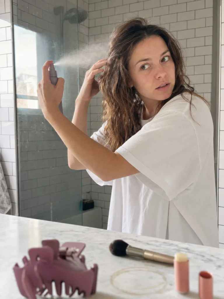 Fair-skinned woman with unstyled medium wavy brown hair holding a texturizing spray bottle mid-spritz in a white-tiled bathroom, with a mauve claw clip resting on a marble vanity in the foreground