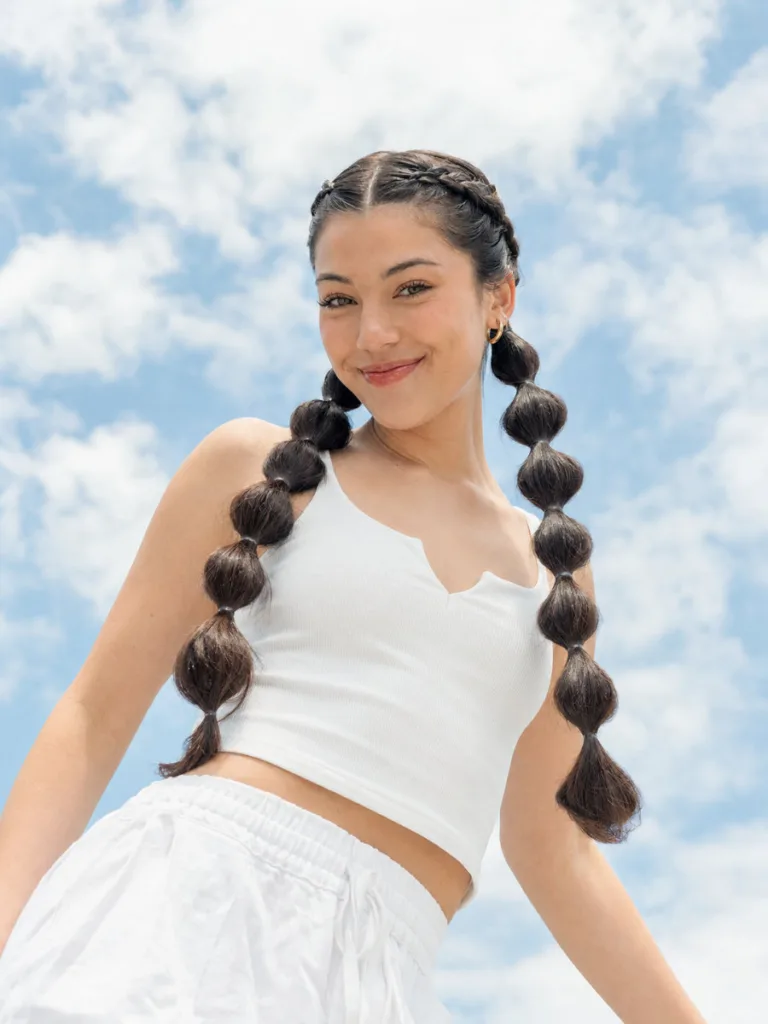 Fair-skinned woman with dutch braid pigtails transitioning into bubble ponytail ends, low-angle shot against blue sky, playful expression