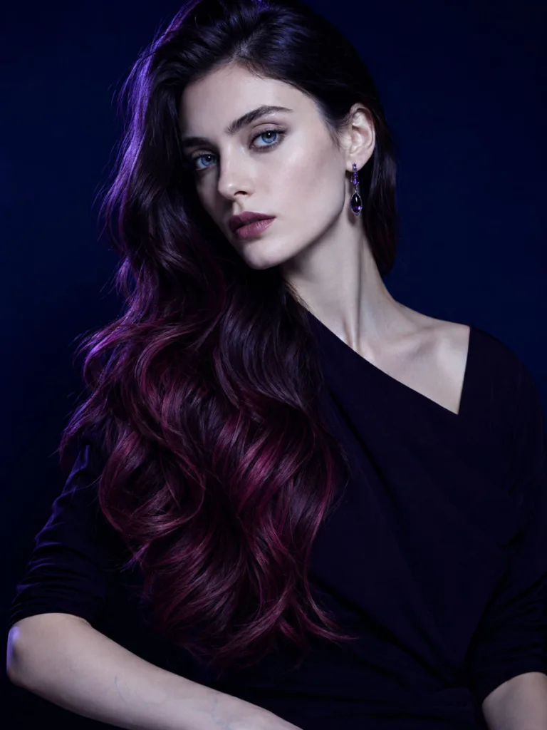 Woman with vivid magenta-burgundy balayage on long dark brown hair swept over one shoulder, midnight blue background, cool editorial lighting
