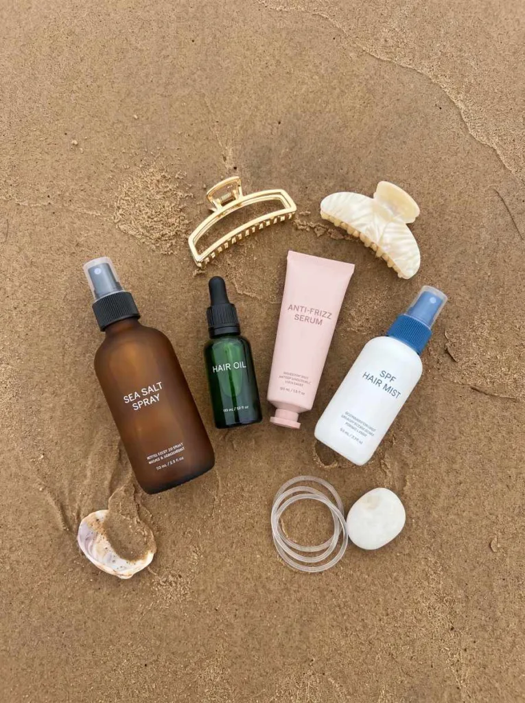 Beach hair essentials flat lay with sea salt spray, anti-frizz serum, UV hair mist, and shell clips on natural sand