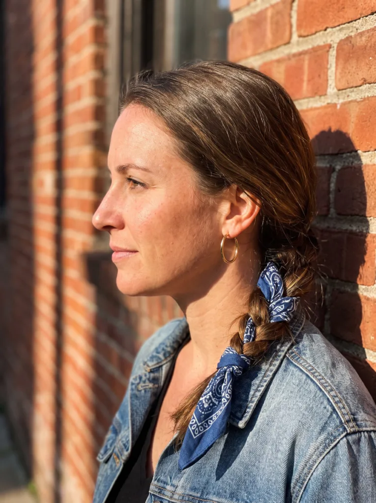 Woman with a low side braid woven with a patterned bandana against an urban brick wall