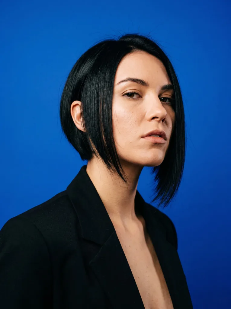 Woman with an asymmetrical bob haircut, one side noticeably longer, photographed against a bold graphic background