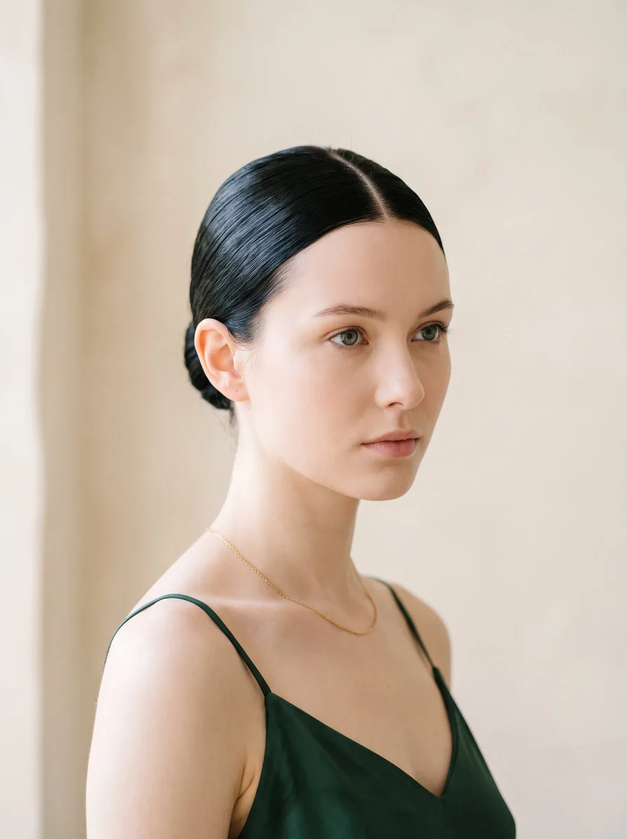 Beautiful 22-year-old woman with porcelain skin and jet black hair in a glass-finish sleek low bun, front-facing portrait, warm cream background