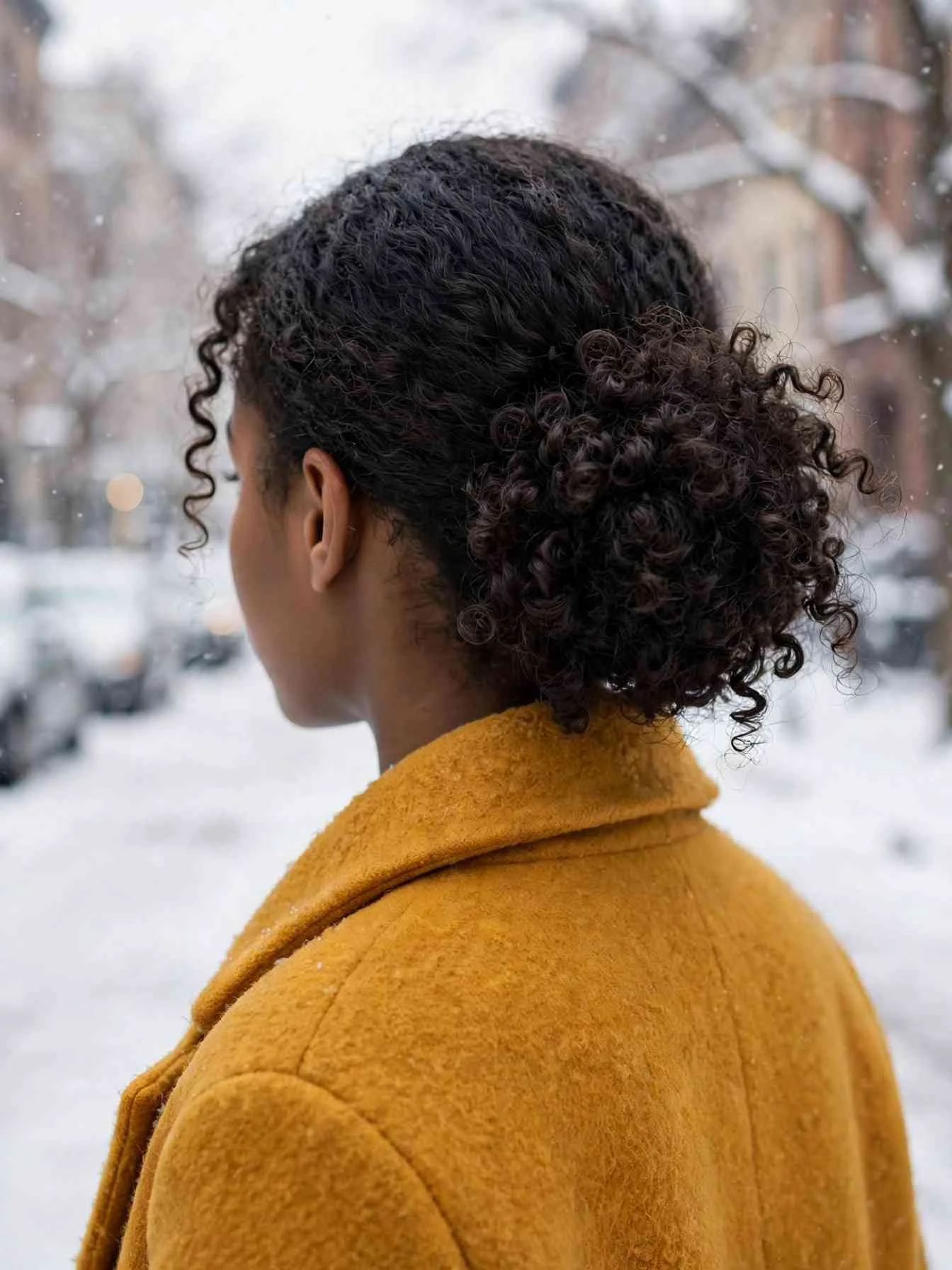 Natural curls pinned into a low rounded full messy bun, mustard wool coat